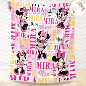 Puede incluir: Manta de beb&eacute; rosa y amarilla con un patr&oacute;n repetido del nombre "Mira" e im&aacute;genes de Mickey Mouse.