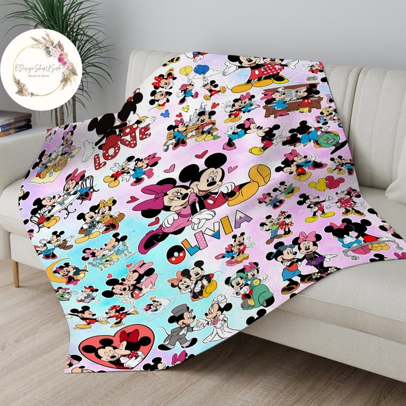 May include: A pink and blue fleece blanket with a repeating pattern of Mickey Mouse and Minnie Mouse characters. The blanket features the names "Love" and "Olivia" in a glittery font.