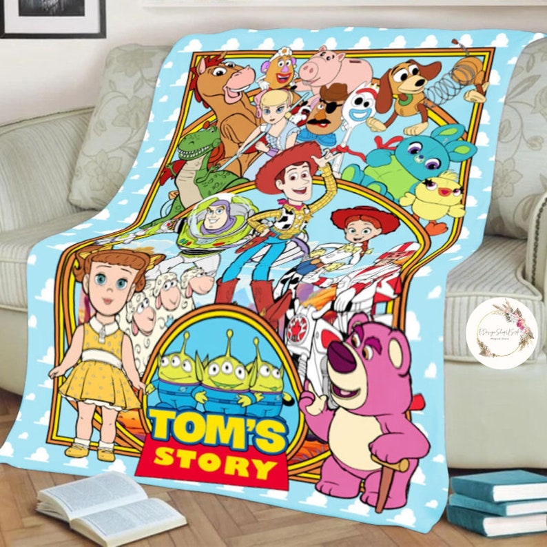 Puede incluir: Una manta colorida con personajes de la franquicia Toy Story. La manta es azul con un borde blanco y presenta personajes como Woody, Buzz Lightyear, Jessie y Bo Peep. El texto "TOM'S STORY" est&aacute; impreso en el centro de la manta.