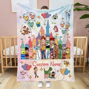 May include: A pink and white fleece blanket with a colorful cartoon image of Toy Story characters. The characters are standing in front of a castle with a pink and white background. The text "Custom Name" is printed on the blanket.