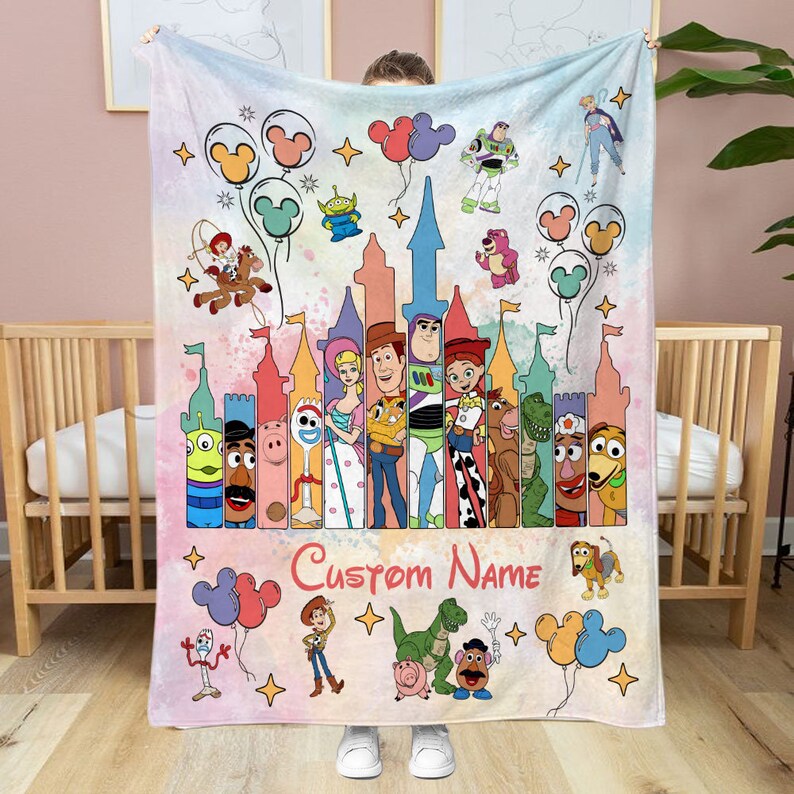 May include: A pink and white fleece blanket with a colorful cartoon image of Toy Story characters. The characters are standing in front of a castle with a pink and white background. The text "Custom Name" is printed on the blanket.
