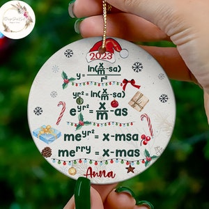 May include: A round white Christmas ornament with a red Santa hat on top. The ornament features a green Christmas tree with lights and presents. The tree is decorated with candy canes and snowflakes. The ornament has a white background with the text "2023" and a math equation that says "merry = x-mas". The ornament is personalized with the name "Anna".