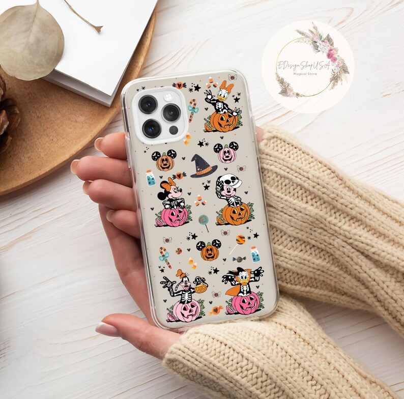 May include: A clear phone case with a Halloween-themed design featuring Mickey Mouse, Minnie Mouse, Donald Duck, and Goofy dressed in costumes. The case has a white background with orange, black, and pink accents. The characters are surrounded by pumpkins, sweets, and stars.