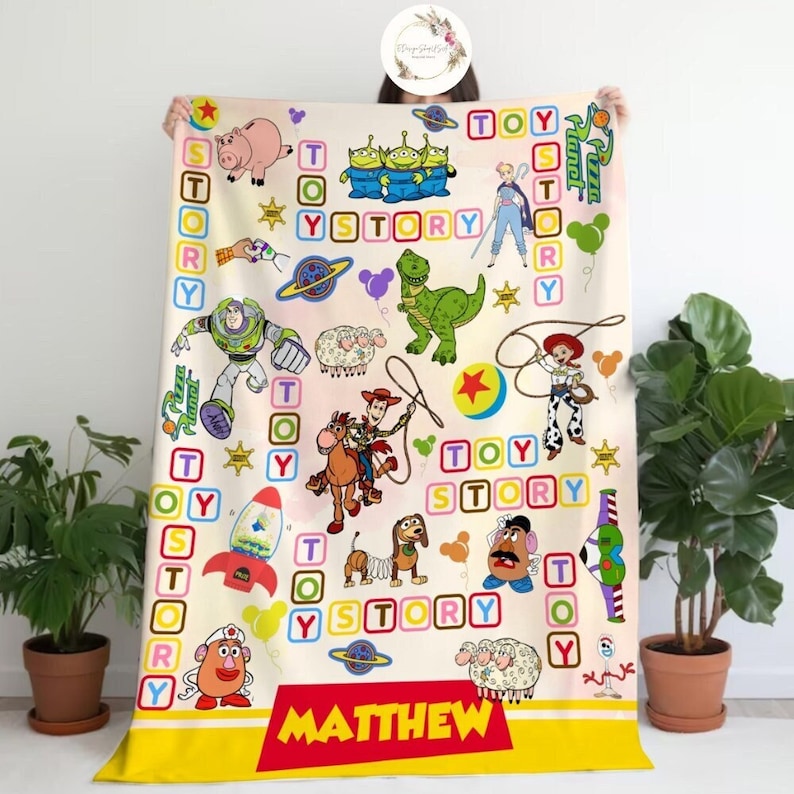 Puede incluir: Una manta amarilla y blanca con tem&aacute;tica de Toy Story. La manta presenta personajes de la pel&iacute;cula, incluidos Woody, Buzz Lightyear y Rex. La manta tambi&eacute;n tiene las palabras "Toy Story" y "Matthew" impresas en ella.