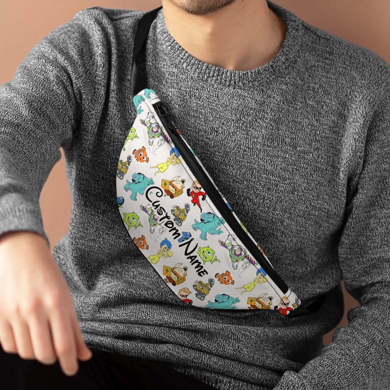 Puede incluir: Ri&ntilde;onera blanca con una correa negra y un estampado colorido de varios personajes de Disney, incluidos Buzz Lightyear, Woody y Sulley. El texto "Custom Name" est&aacute; impreso en la ri&ntilde;onera.