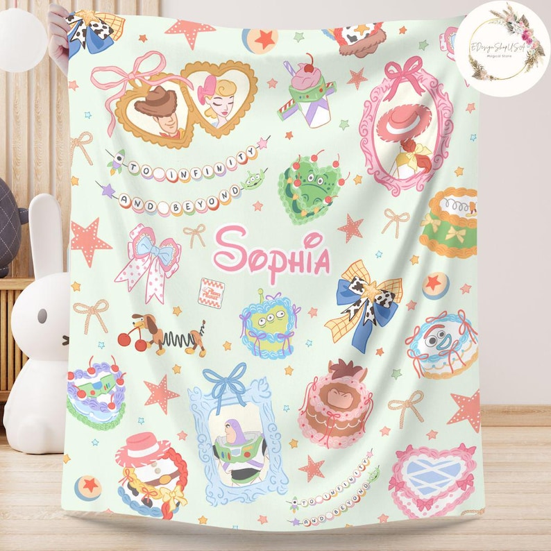 May include: A green and pink baby blanket with a Toy Story theme. The blanket features characters from the movie, including Woody, Buzz Lightyear, and Jessie. The blanket also has a personalized name, "Sophia", in a pink font.