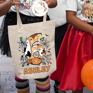 Puede incluir: Una bolsa de lona beige con un dise&ntilde;o de tem&aacute;tica de Halloween que presenta el n&uacute;mero 6, una telara&ntilde;a y personajes de la franquicia Toy Story. La bolsa tiene el nombre "ASHLEY" impreso en la parte delantera.