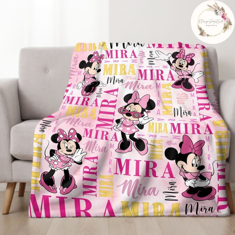 Puede incluir: Manta de forro polar rosa con un patr&oacute;n repetido del nombre "Mira" en letras rosas y amarillas. La manta presenta cuatro im&aacute;genes de Minnie Mouse en diferentes poses, con un vestido rosa con lunares blancos y un lazo rojo.