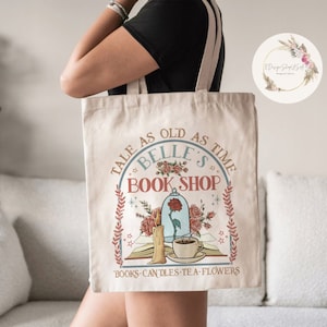 May include: A beige tote bag with a design featuring the text "Tale as old as time Belle's Book Shop" with a floral design and a glass dome with a rose inside. The text "Books Candles Tea Flowers" is at the bottom of the design.