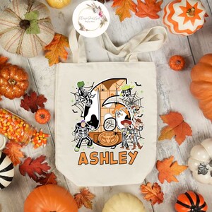 Puede incluir: Una bolsa de lona blanca con un dise&ntilde;o de tem&aacute;tica de Halloween con los personajes de Toy Story vestidos de esqueletos. El n&uacute;mero "6" est&aacute; en el fondo con el nombre "ASHLEY" impreso debajo.