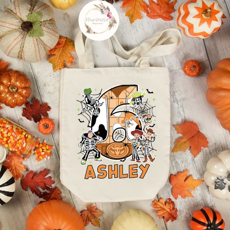 Puede incluir: Una bolsa de lona blanca con un dise&ntilde;o de tem&aacute;tica de Halloween con los personajes de Toy Story vestidos de esqueletos. El n&uacute;mero "6" est&aacute; en el fondo con el nombre "ASHLEY" impreso debajo.