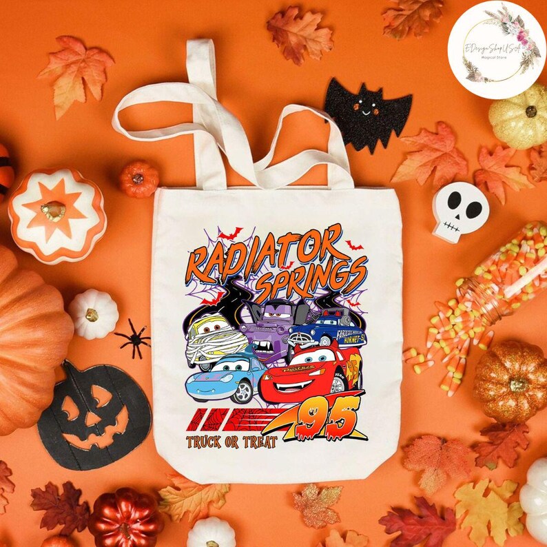 Op de afbeelding: Een witte canvas draagtas met een Halloween-ontwerp met Cars-personages verkleed als mummies en vampiers, met de tekst "Radiator Springs" en "Truck or Treat". De tas is omgeven door pompoenen, bladeren en Halloween-decoraties.