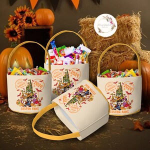 May include: Three Halloween treat bags with a Mickey Mouse theme. The bags are beige with a yellow handle and feature a print of Mickey Mouse and friends in front of Cinderella's Castle. The bags are filled with candy and have the text "Mickey's Halloween Party 2024" printed on them.