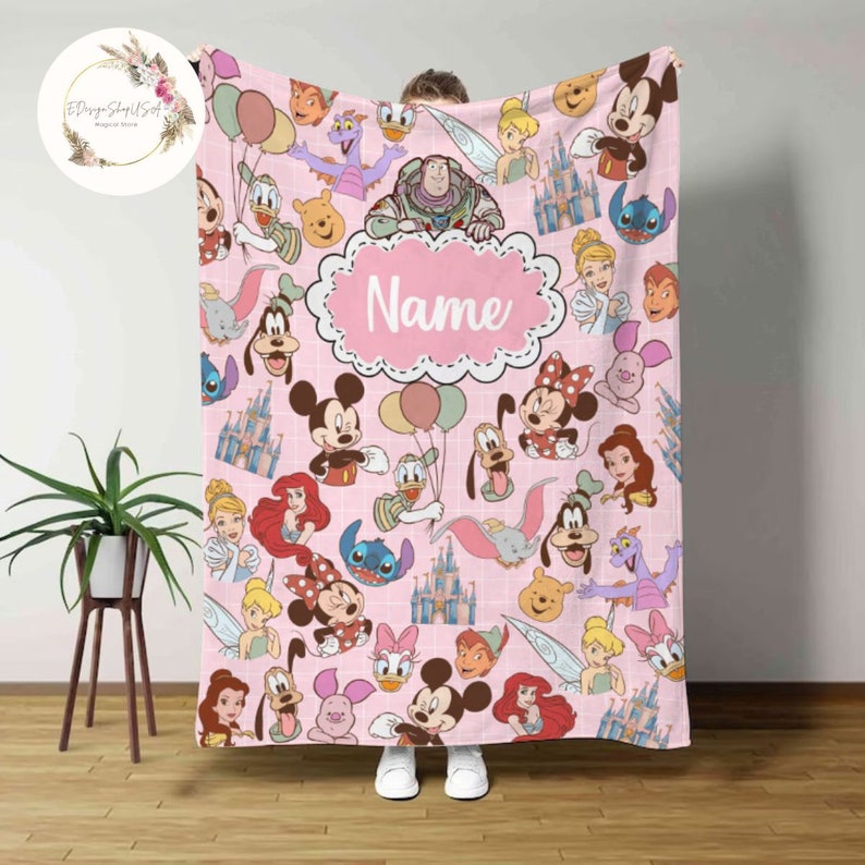 May include: Pink fleece blanket with a pattern of various Disney characters, including Mickey Mouse, Minnie Mouse, Donald Duck, Goofy, and more. The blanket has a white grid pattern and a pink oval with the word "Name" in white text.
