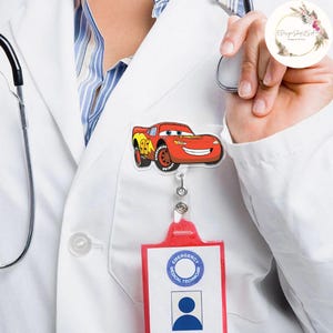 Puede incluir: Un carrete de insignia de Rayo McQueen rojo y amarillo con una cara sonriente, adjunto a un soporte de identificaci&oacute;n rojo. El soporte de identificaci&oacute;n tiene las palabras "T&eacute;cnico M&eacute;dico de Emergencias" y una silueta de una persona.