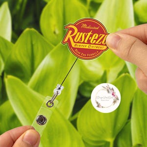 Puede incluir: Un portatarjetas retr&aacute;ctil con un logotipo redondo rojo y amarillo que dice "Rust-eze Bumper Ointment Rear End Formula". El portatarjetas es sostenido por una mano contra un fondo de hojas verdes. El portatarjetas tiene un soporte de pl&aacute;stico transparente.