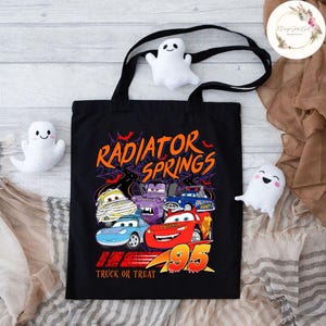 Op de afbeelding: Zwarte draagtas met een kleurrijke afbeelding van auto's uit Radiator Springs, met de tekst "Radiator Springs" en "Truck or Treat". De tas heeft zwarte handvatten en is versierd met drie kleine witte spookknuffels.