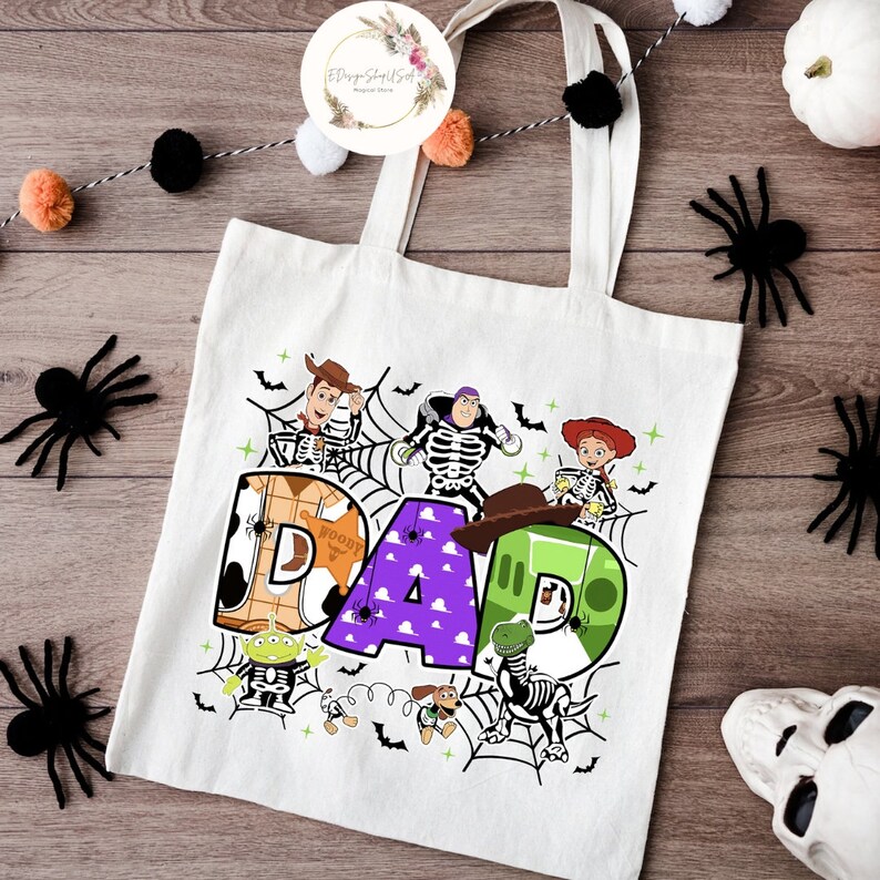 Puede incluir: Bolsa de tela blanca con un dise&ntilde;o colorido con personajes de la pel&iacute;cula Toy Story. El dise&ntilde;o incluye la palabra "DAD" con los personajes vestidos de esqueletos. Los personajes incluyen a Woody, Buzz Lightyear, Jessie, Rex y Slinky.