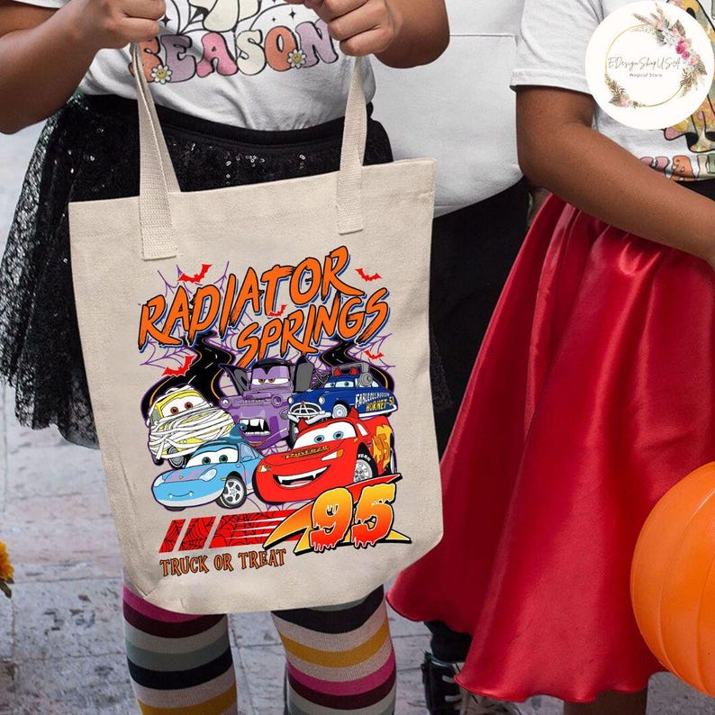 Op de afbeelding: Beige canvas draagtas met een Halloween-thema. Het ontwerp toont cartoonauto's in kostuums en de tekst "Radiator Springs" en "Truck or Treat". De tas heeft twee handvatten.