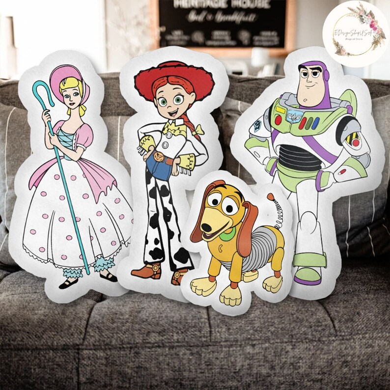 Op de afbeelding: Vier pluche kussens met personages uit de Toy Story-franchise. De kussens zijn wit met kleurrijke cartoonafbeeldingen van Woody, Buzz Lightyear, Jessie en Slinky.
