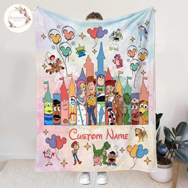 May include: A colorful fleece blanket with a watercolor background featuring a Disney castle and characters from the Toy Story franchise. The castle is surrounded by Mickey Mouse balloons and stars. The text "Custom Name" is printed below the characters.