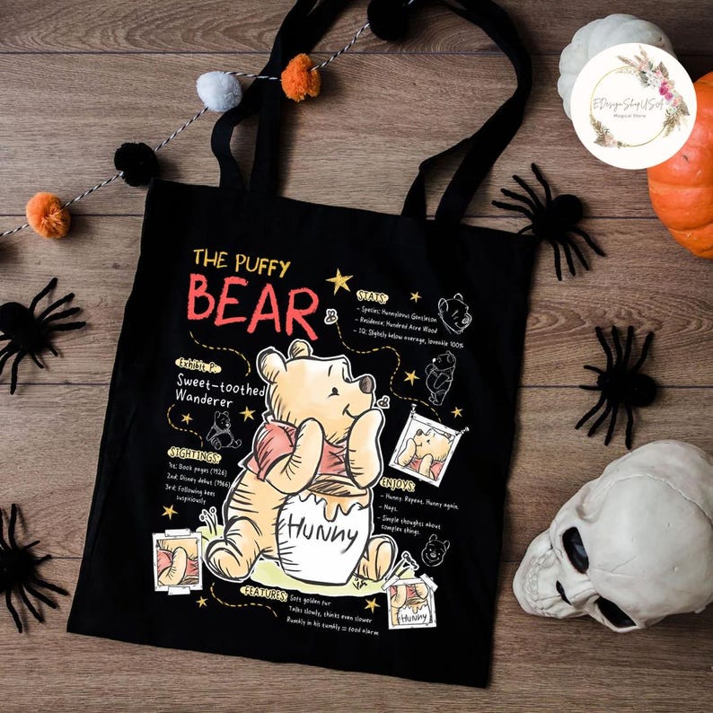 May include: Black tote bag featuring Winnie the Pooh with the text "The Puffy Bear". The design includes illustrations and text about Pooh's characteristics and interests. The bag has black handles and is decorated with Halloween-themed items.