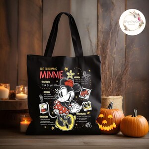 May include: Black tote bag featuring Minnie Mouse with the text "The Charming Minnie, The Style Icon." The design includes illustrations and text about Minnie. Two carved pumpkins and candles are in the background.