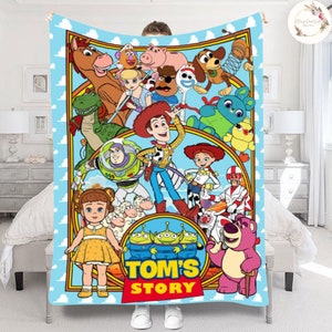 Puede incluir: Una manta colorida con personajes de la franquicia Toy Story, incluyendo Woody, Buzz Lightyear, Jessie y los Aliens. La manta tiene un fondo azul con nubes blancas y un borde amarillo. El texto "TOM'S STORY" est&aacute; impreso en el centro de la manta.