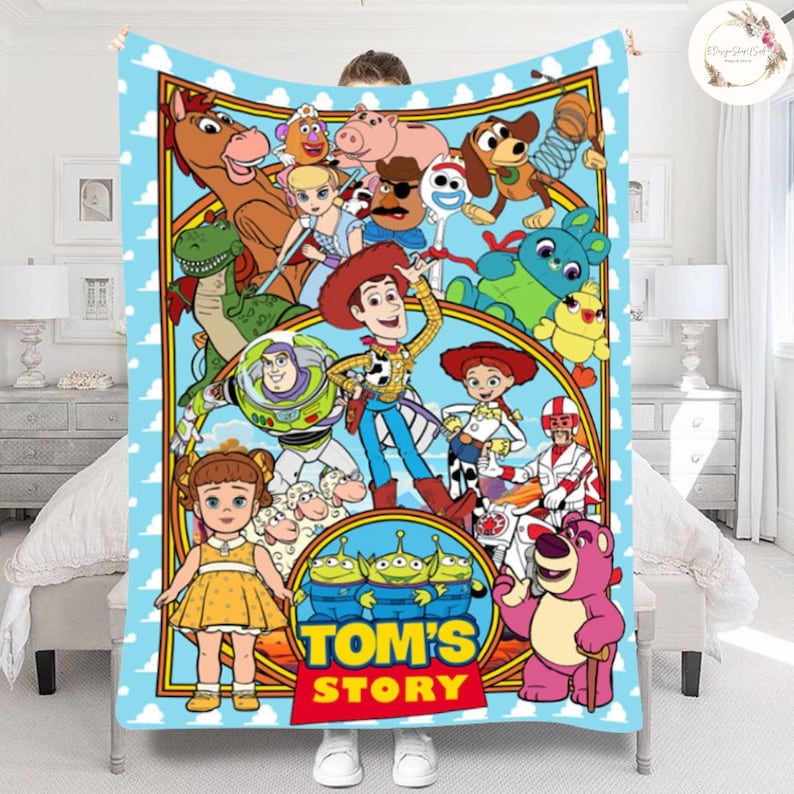 Puede incluir: Una manta colorida con personajes de la franquicia Toy Story, incluyendo Woody, Buzz Lightyear, Jessie y los Aliens. La manta tiene un fondo azul con nubes blancas y un borde amarillo. El texto "TOM'S STORY" est&aacute; impreso en el centro de la manta.