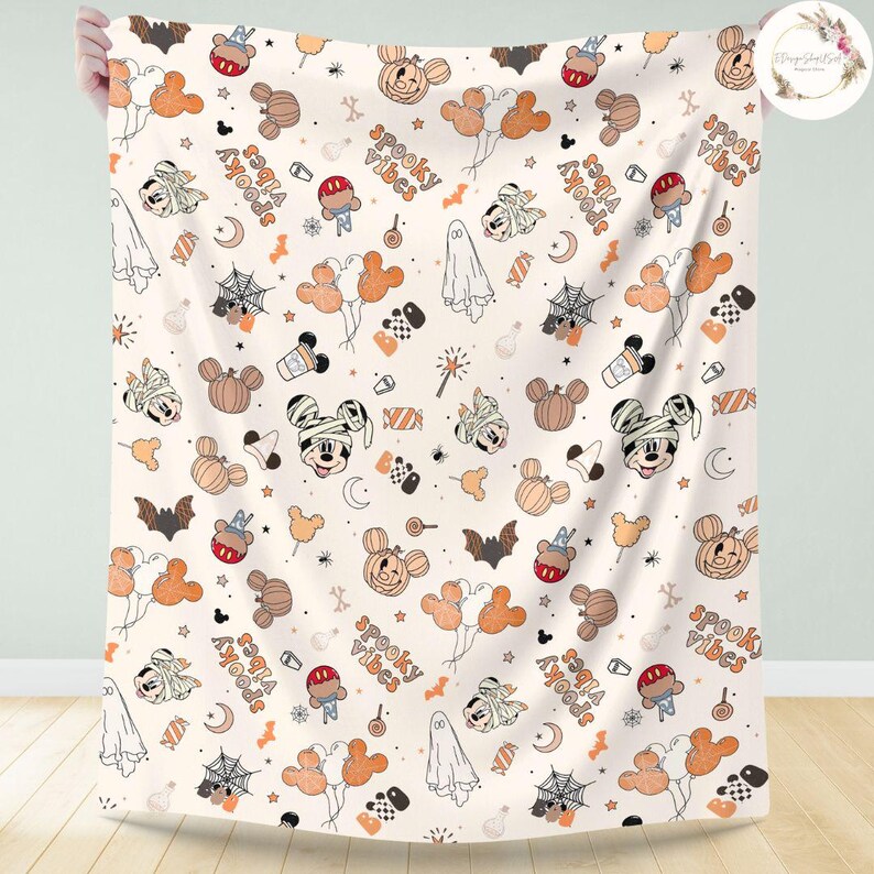 May include: A white fabric blanket with a Halloween-themed pattern featuring Mickey Mouse in a mummy costume, pumpkins, bats, ghosts, and the text "spooky vibes".