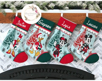 May include: Four personalized Christmas stockings with a blue and green background. Each stocking features a different Disney character: Donald Duck, Mickey Mouse, Minnie Mouse, and Goofy. The stockings are hung on a wooden mantel.