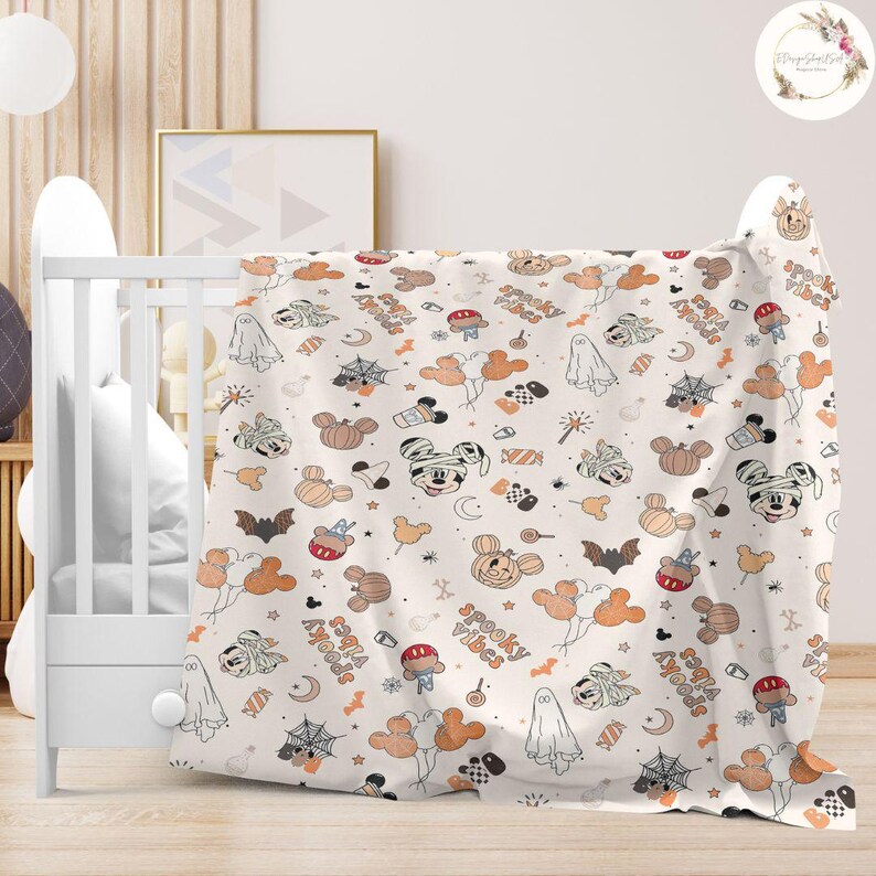 May include: A white baby blanket with a Halloween-themed pattern featuring Mickey Mouse and other Disney characters. The pattern includes pumpkins, ghosts, bats, candy corn, and the text "Spooky Vibes" and "Sweet Treats".