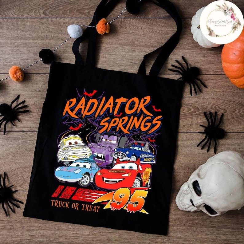 Op de afbeelding: Zwarte draagtas met een afbeelding van Cars-personages, de tekst "Radiator Springs" en "Truck or Treat". Het ontwerp bevat het nummer 95 in oranje. Halloween-decoraties, waaronder spinnen en een schedel, omringen de tas.