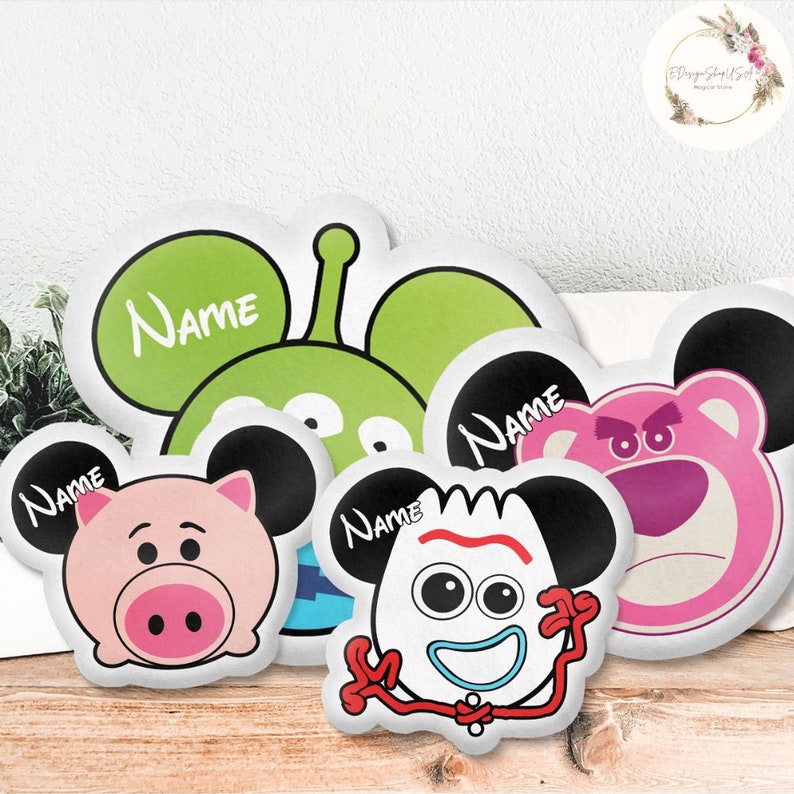 K&ouml;nnte beinhalten: F&uuml;nf wei&szlig;e Pl&uuml;schkissen mit verschiedenen Comicfiguren. Jedes Kissen hat schwarze Mickey-Maus-Ohren und das Wort "NAME" auf der Vorderseite aufgedruckt. Die Figuren sind ein gr&uuml;ner Au&szlig;erirdischer, ein rosa Schwein, eine blaue und wei&szlig;e Figur, eine wei&szlig;e Figur mit einem roten Streifen und ein rosa B&auml;r.