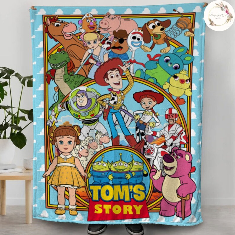 Puede incluir: Una manta colorida con personajes de la franquicia de pel&iacute;culas Toy Story. La manta presenta a Woody, Buzz Lightyear, Jessie y otros personajes de la pel&iacute;cula. La manta es azul con un borde blanco y tiene las palabras "Tom's Story" en letras rojas.