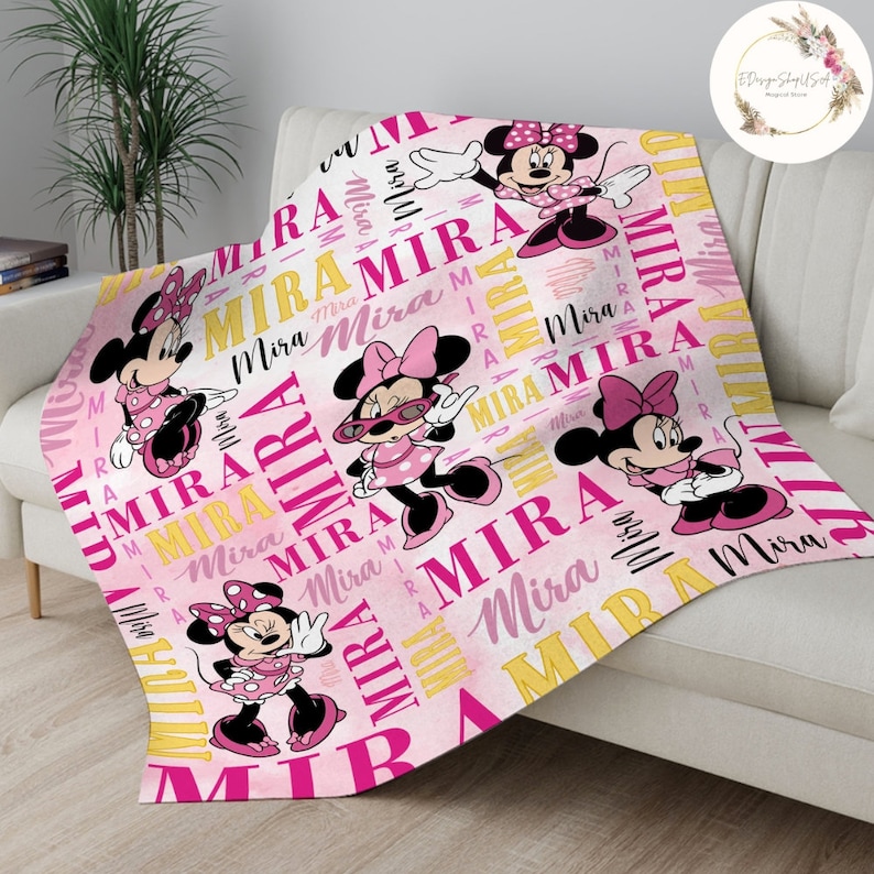 Puede incluir: Manta personalizada rosa y amarilla con un patr&oacute;n repetido del nombre "Mira" e im&aacute;genes de Mickey Mouse con un vestido rosa.