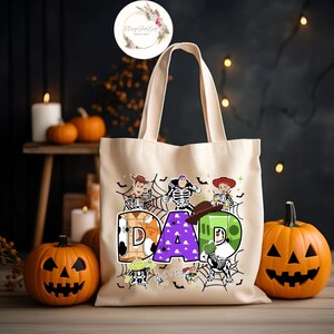 Puede incluir: Una bolsa de lona beige con un dise&ntilde;o de tem&aacute;tica de Halloween con los personajes de Toy Story vestidos de esqueletos. El dise&ntilde;o incluye la palabra "DAD" con cada letra decorada con un personaje diferente y elementos de Halloween como murci&eacute;lagos y telara&ntilde;as.