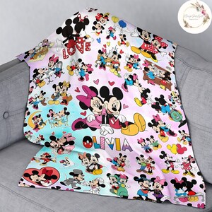 May include: A pink and blue blanket with a repeating pattern of Mickey Mouse and Minnie Mouse. The blanket features a variety of images of the characters, including them holding hands, playing music, and kissing. The blanket also features the word "LOVE" and the name "OLIVIA".