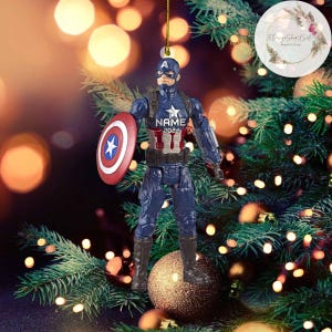K&ouml;nnte beinhalten: Ein rot-wei&szlig;-blauer Captain America Weihnachtsbaumschmuck mit einem Schild und dem Text "NAME 2024" auf der Vorderseite.