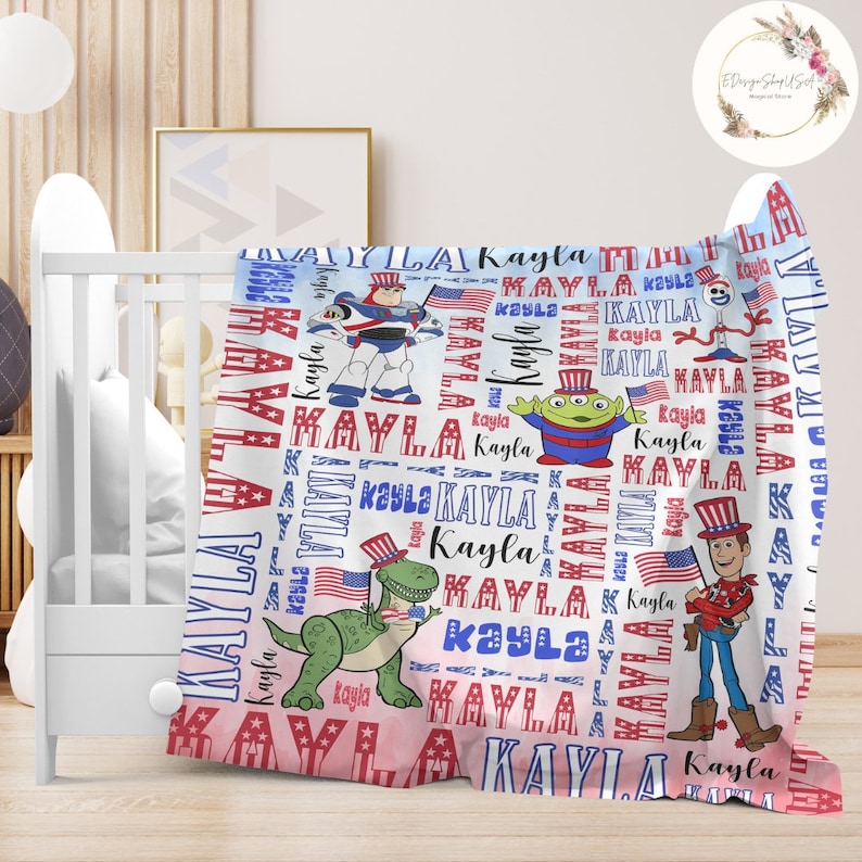Puede incluir: Una manta de beb&eacute; blanca y rosa con un tema patri&oacute;tico rojo, blanco y azul. La manta presenta un patr&oacute;n repetido del nombre "Kayla" e im&aacute;genes de personajes de Toy Story con sombreros patri&oacute;ticos. Los personajes incluyen Buzz Lightyear, Woody y el extraterrestre.