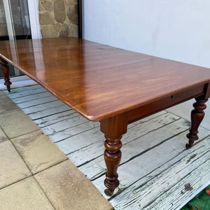 May include: A large, rectangular, wooden dining table with a dark brown finish. The table has a drop leaf on each side and sits on four turned legs with decorative details. The table is shown on a whitewashed wooden floor.