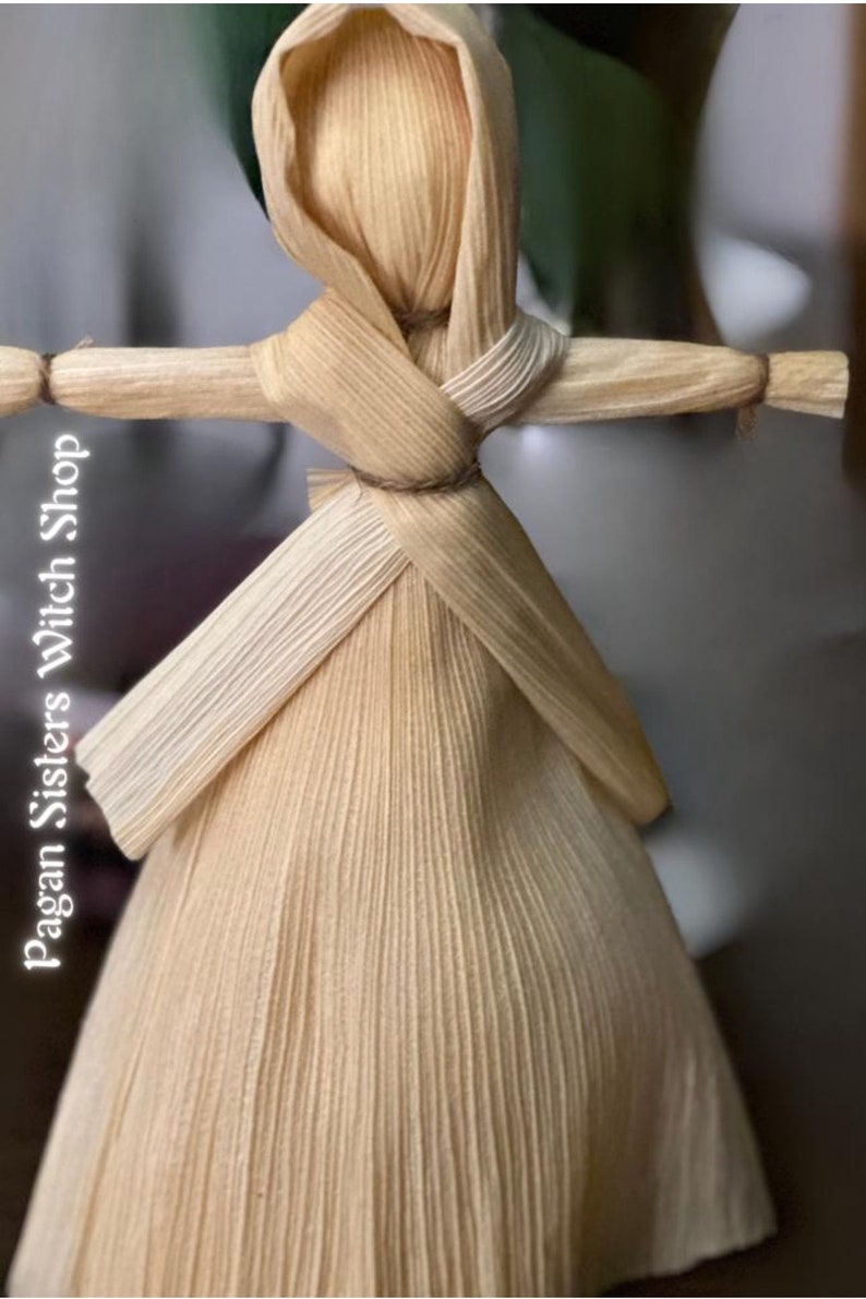 May include: A handmade corn husk doll with a long, flowing dress and a hood. The doll is tied with twine at the waist and has a simple, rustic look.