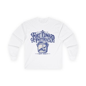 May include: A white long-sleeve shirt featuring a blue graphic print on the chest. The design displays the text The Fort Edward Pottery Co. above an illustration of a ceramic crock with the number 3 and a bird, with Est. 1859 below it.