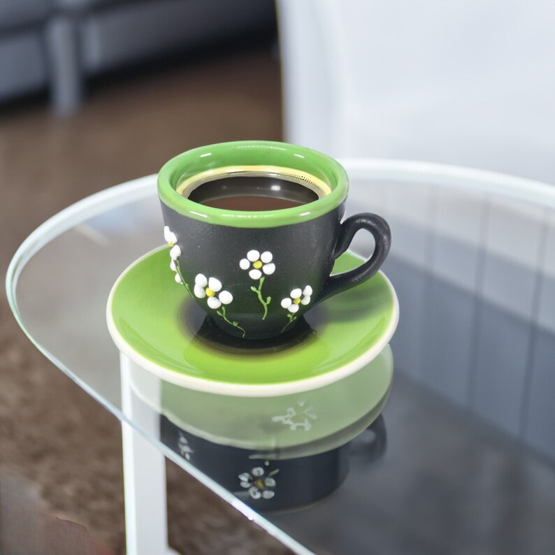 May include: A black and green coffee cup with white flower designs sits on a green saucer. The cup is filled with coffee.