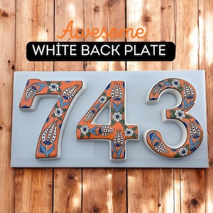 May include: A white ceramic house number sign with the numbers 743 in orange and blue floral designs. The sign is mounted on a wooden surface.
