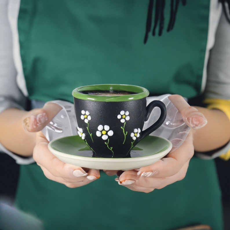 May include: A black coffee cup with a green rim and white daisy designs is being held by a person wearing a green apron. The cup is sitting on a green saucer.