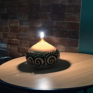 Può includere: Una lampada ad olio in ceramica bianca con un design a spirale bianco e nero. La lampada è accesa e proietta un bagliore caldo.