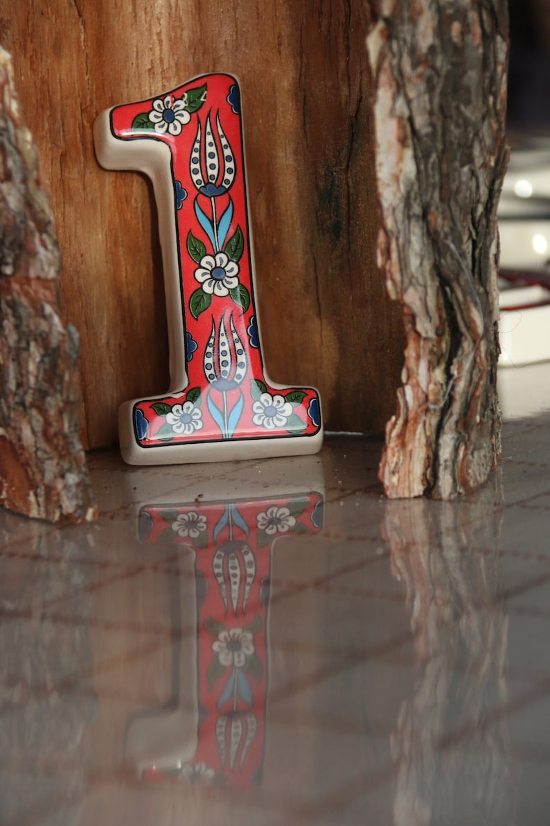 May include: A red ceramic number one with a white background and blue and green floral designs. The number is sitting on a wooden surface.