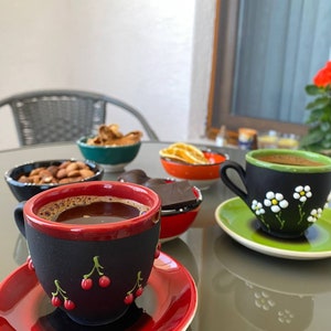 May include: A table setting with a variety of snacks and drinks, including a cup of coffee with a black and red cherry design, a cup of tea with a black and green floral design, a bowl of nuts, a bowl of dried fruit, and a bowl of chocolate.
