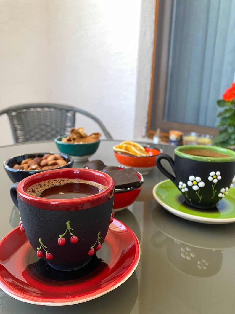 May include: A table setting with a variety of snacks and drinks, including a cup of coffee with a black and red cherry design, a cup of tea with a black and green floral design, a bowl of nuts, a bowl of dried fruit, and a bowl of chocolate.